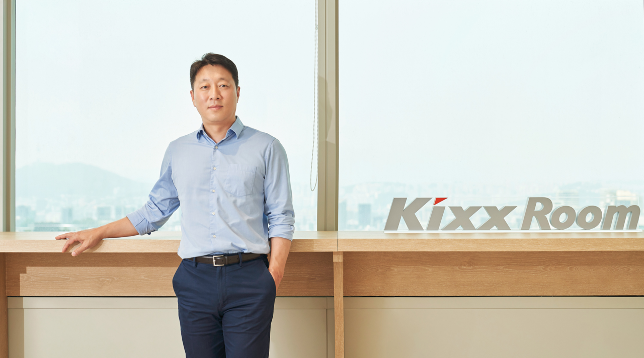 Smart Slider | Kixx Newsroom Kim Hyun Wook, dressed in business casual, looks into the camera as he stands next to a sign reading “Kixx Room.”