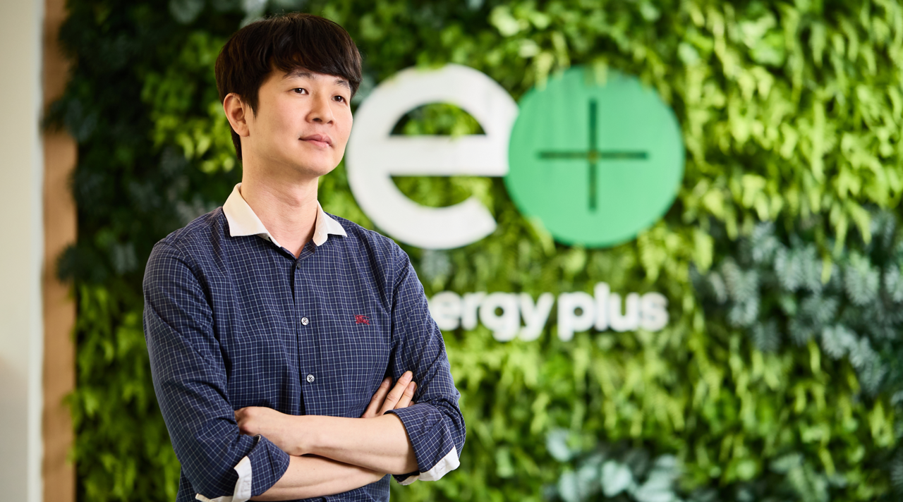 thumbnail | Kixx Newsroom Kixx manager Kwan Hee Choi wears a blue button-up shirt and crosses his arms in front of a leafy wall with the sign “energy plus.”