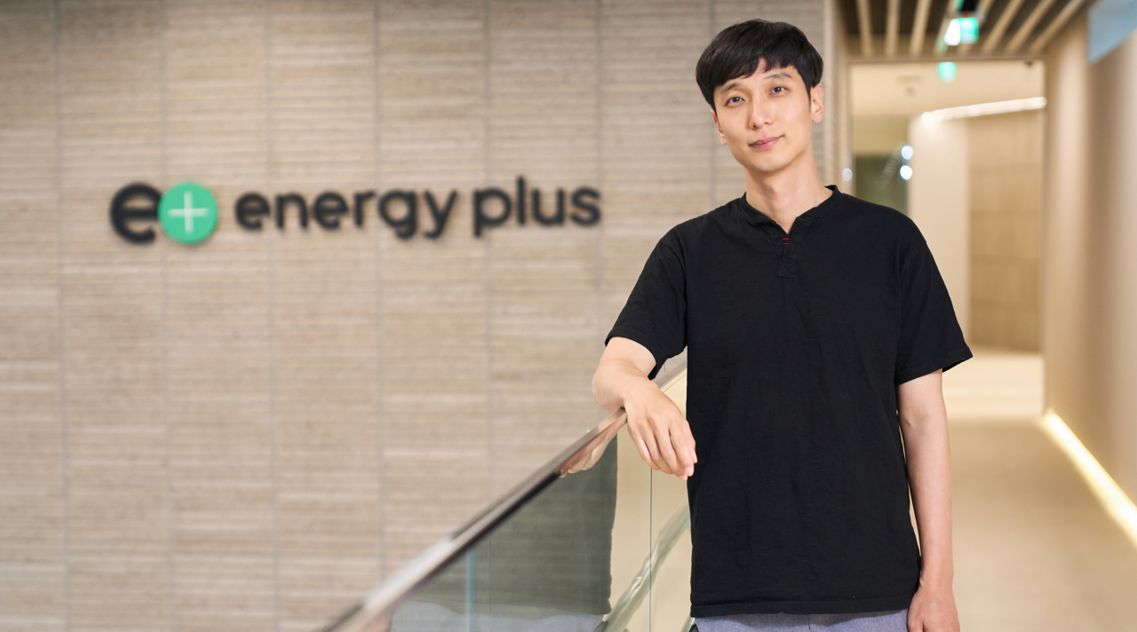 Image 1 | Kixx Newsroom Rae Wook Cho, dressed casually, looks into the camera as he stands with his right arm resting on a railing, in front of “Energy Plus” signage.
