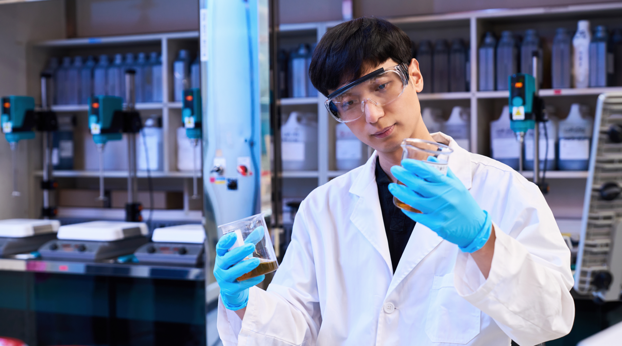 Kixx 2_2 | Kixx Newsroom Rae Wook Cho in a laboratory, dressed in a white lab coat and wearing rubber gloves and safety goggles, examining substances in beakers.