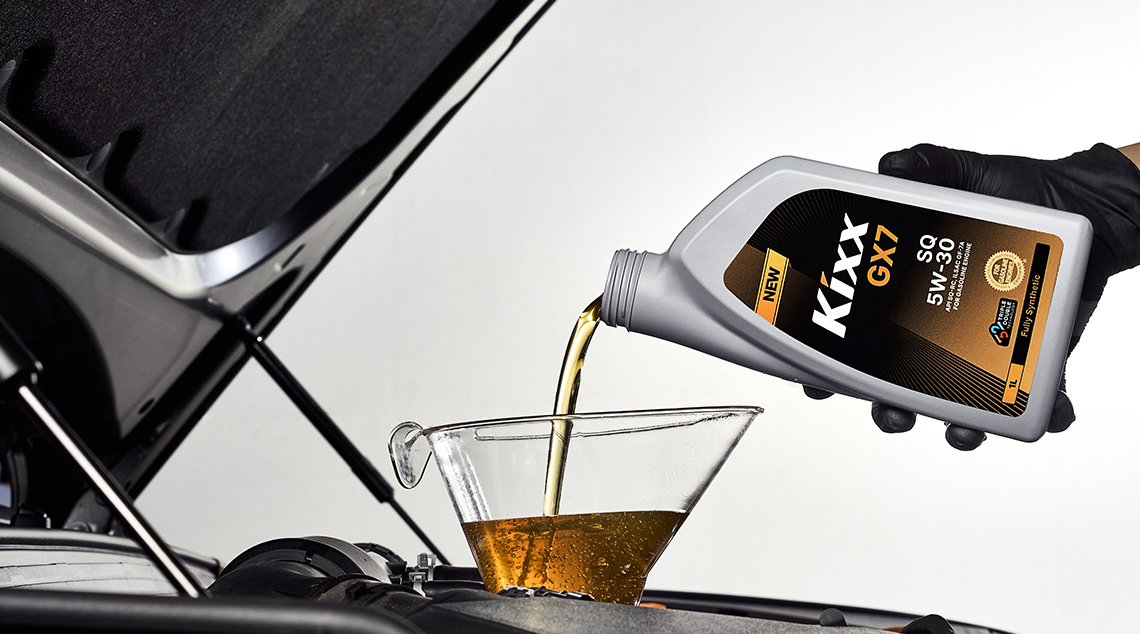 A closeup of a gloved hand pouring a bottle of Kixx GX7 into a car’s engine. 