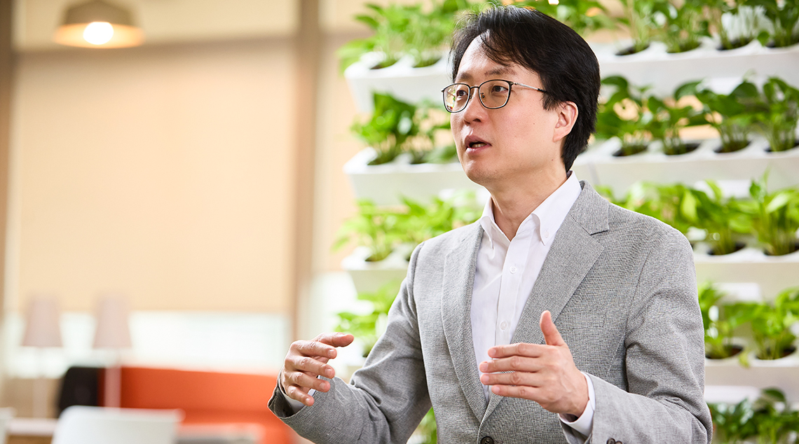 Image 3 | Kixx Newsroom Changbeom Lee wears a grey suit and stands as he speaks animatedly, his arms raised in a gestural pose in a room filled with plants.