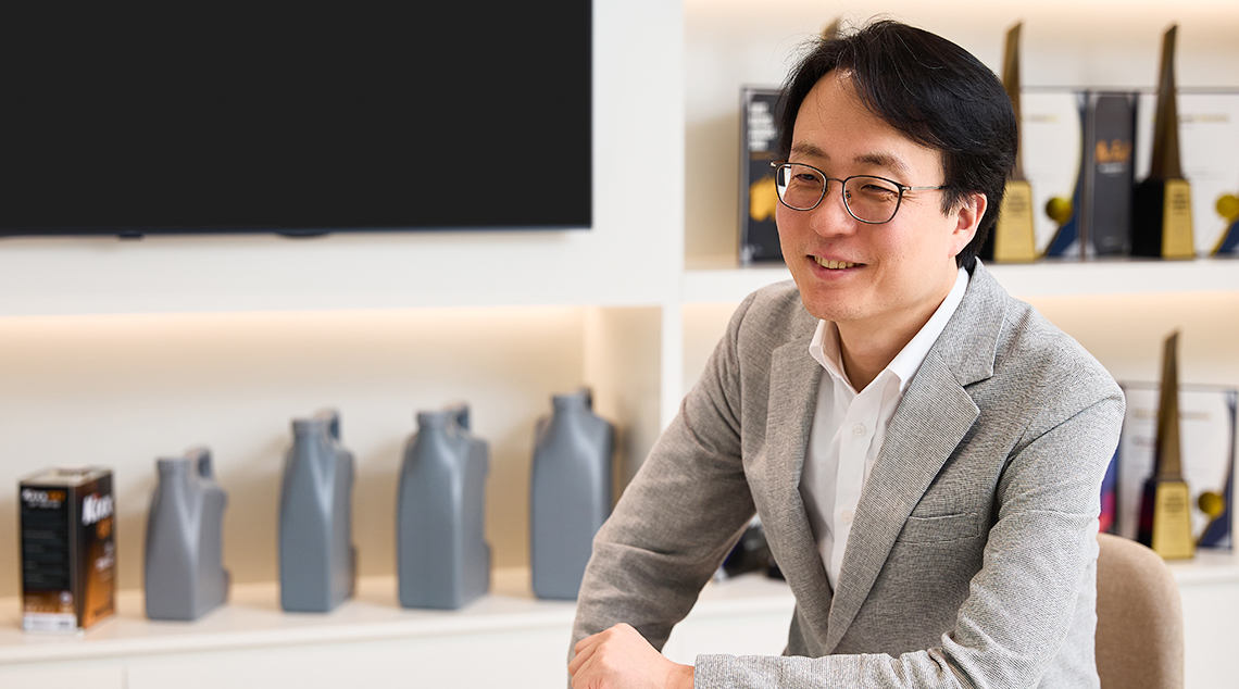 Image 2 | Kixx Newsroom Changbeom Lee smiles and wears a grey suit, sitting with an arm placed on a small table in a room filled with Kixx products and awards.