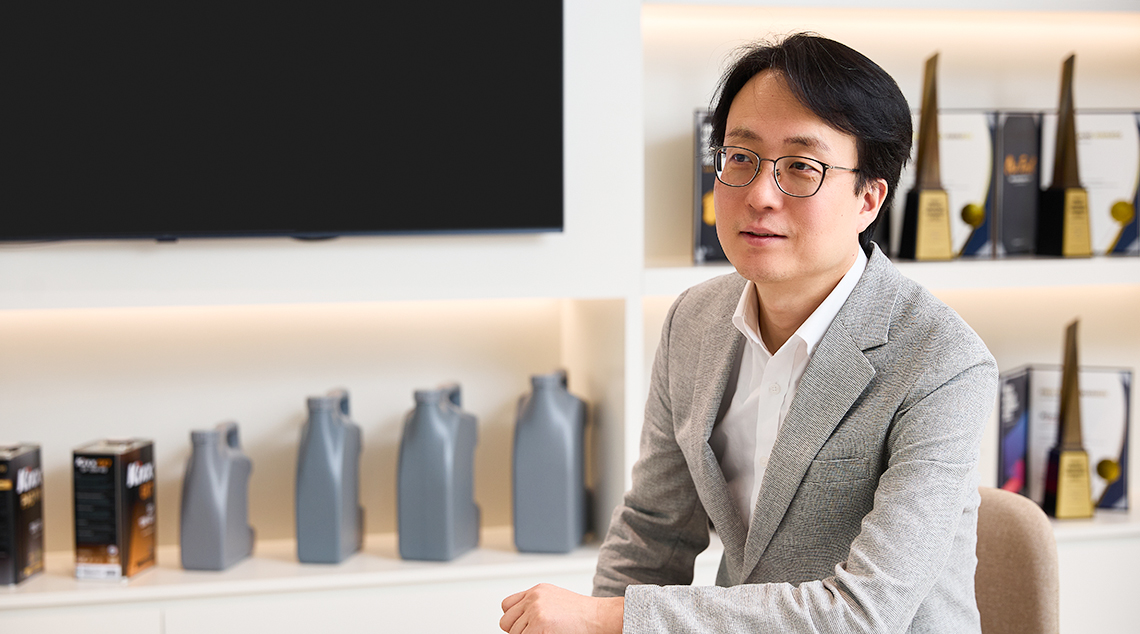 Image 1 | Kixx Newsroom Changbeom Lee wears a grey suit and sits with an arm placed on a small table in a room filled with Kixx products and awards.